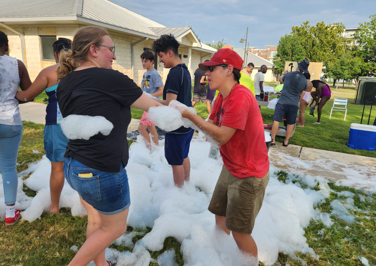 An adult dances with an adolescent outdoors with blasts of foam on the ground and in their hands. A group splashes in the foam.