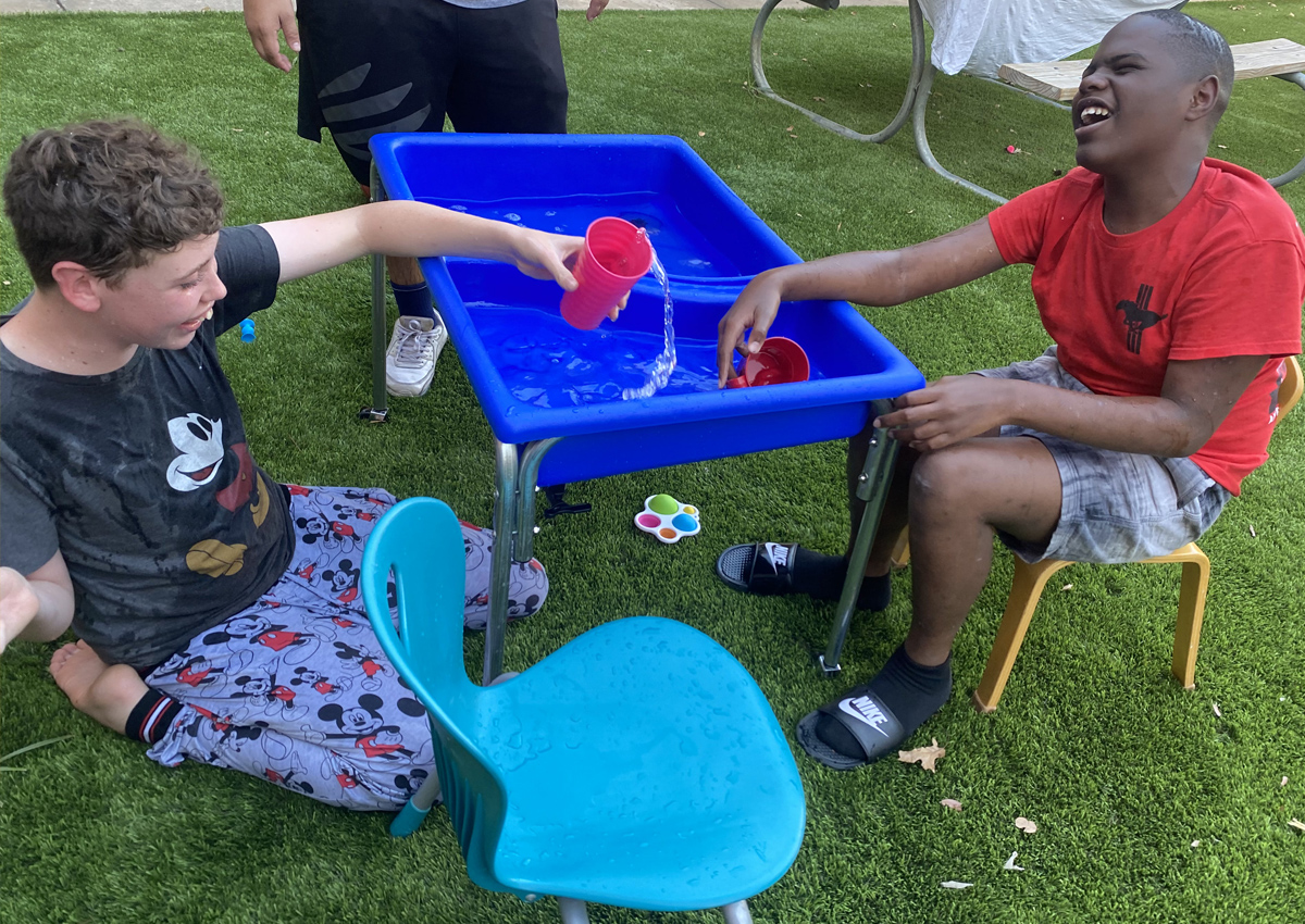 Two adolescents laugh as they scoop water from a large water tray outdoors and splash it on each other.