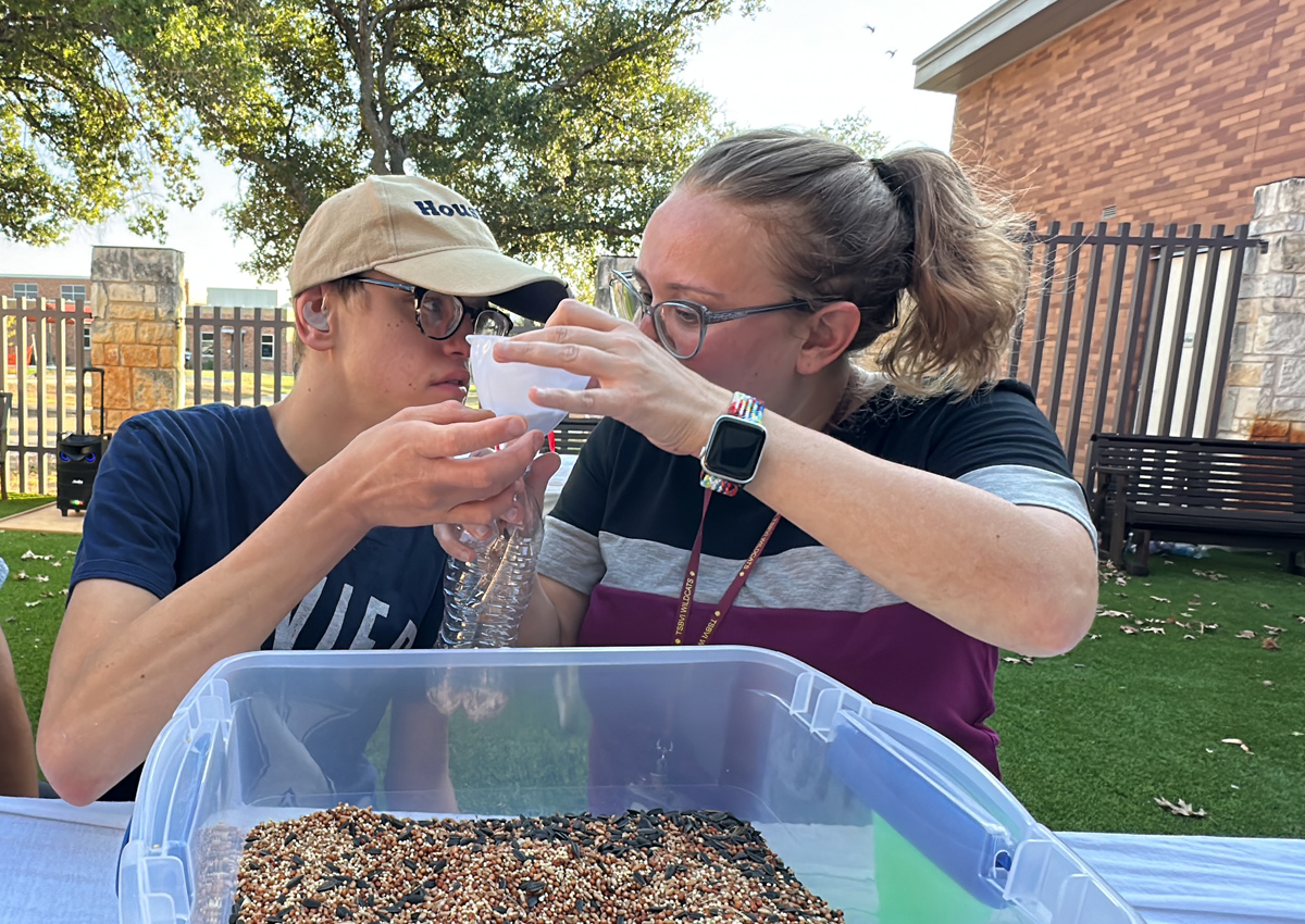 An adult and adolescent seated at a picnic table outdoors pour bird seed into a plastic container using a funnel.