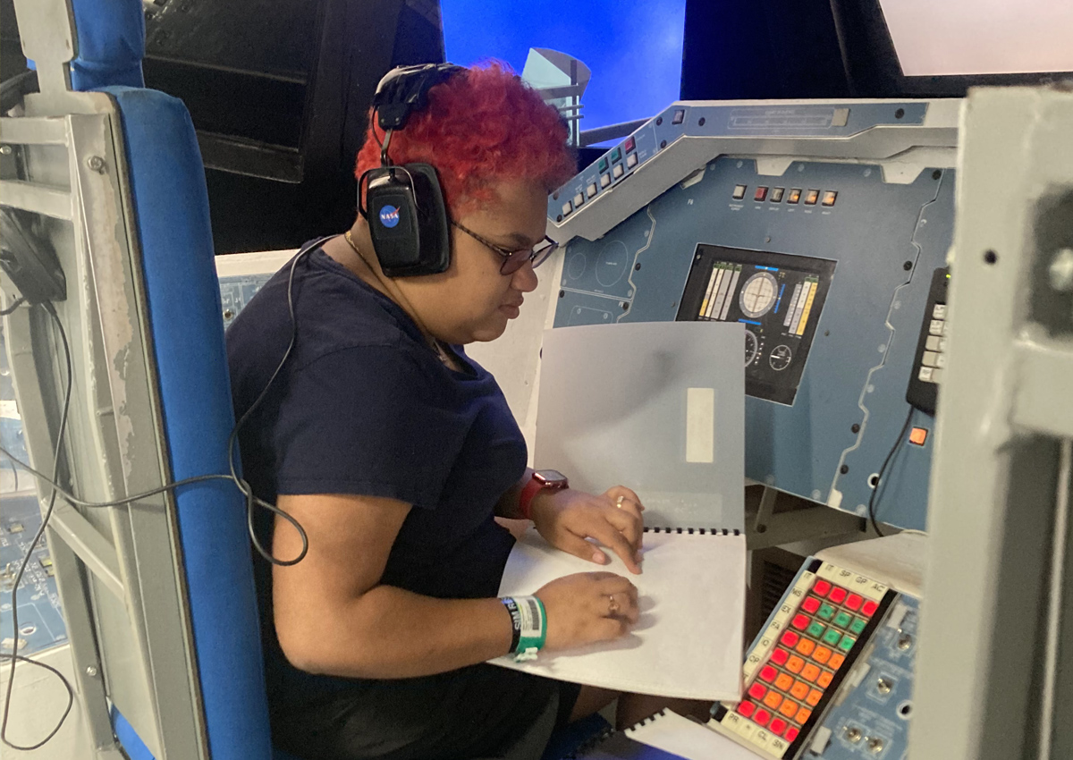 An adolescent reads from a Braille book on her lap in the cockpit of a space shuttle.