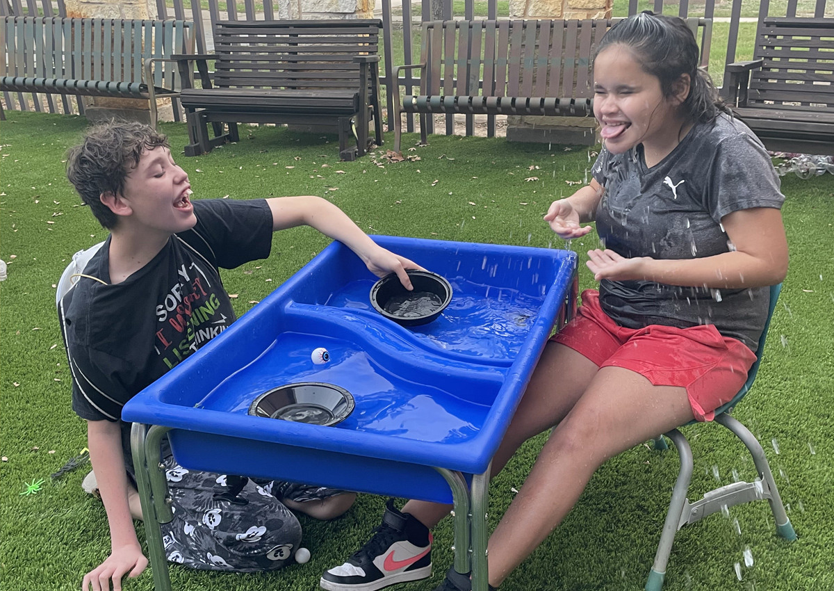 Two adolescents seated at a large water table outdoors splash themselves.