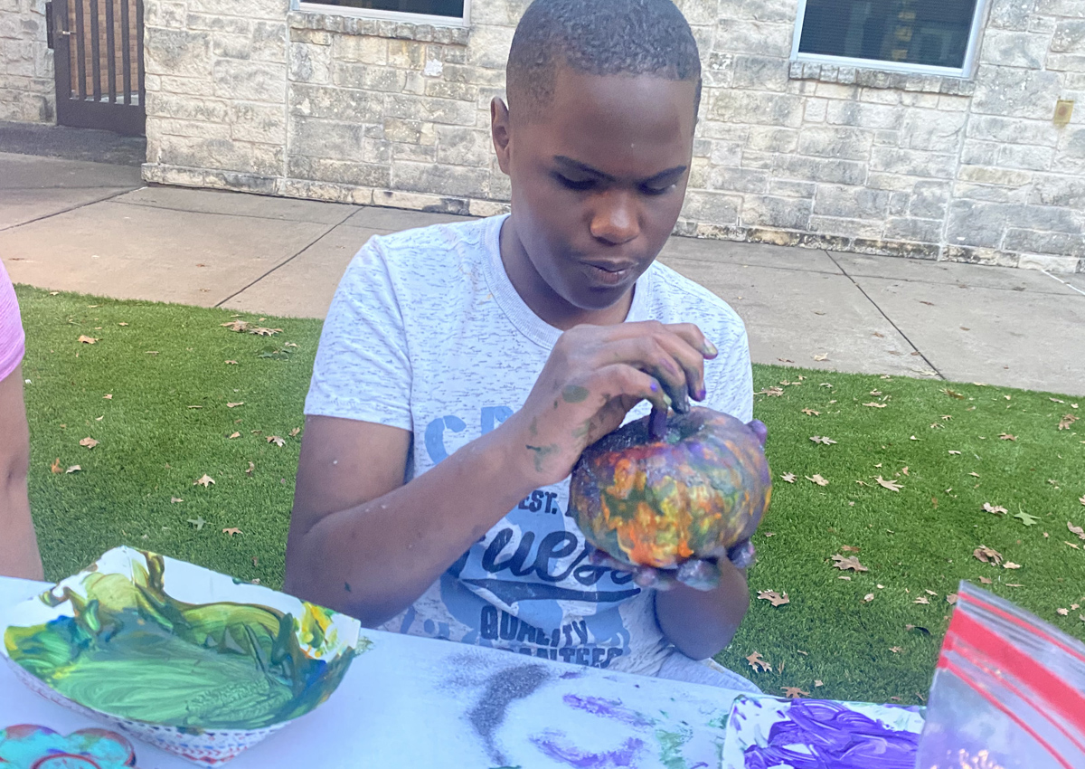 An adolescent seated at a picnic table outdoors paints a pumpkin on a table filled with paint, glitter, and crafts.