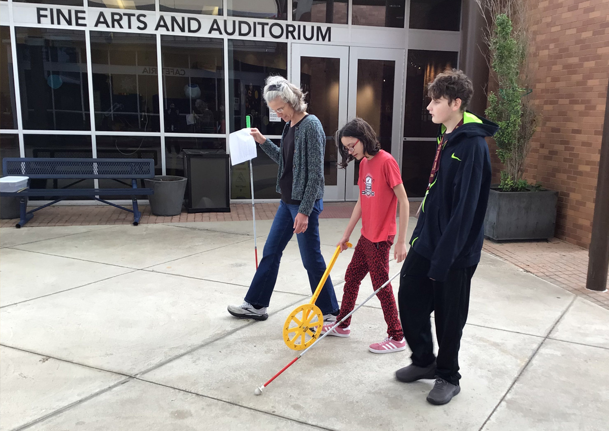 An adult and two adolescents walk outside. One uses a trundle wheel and the other walks with a white cane.