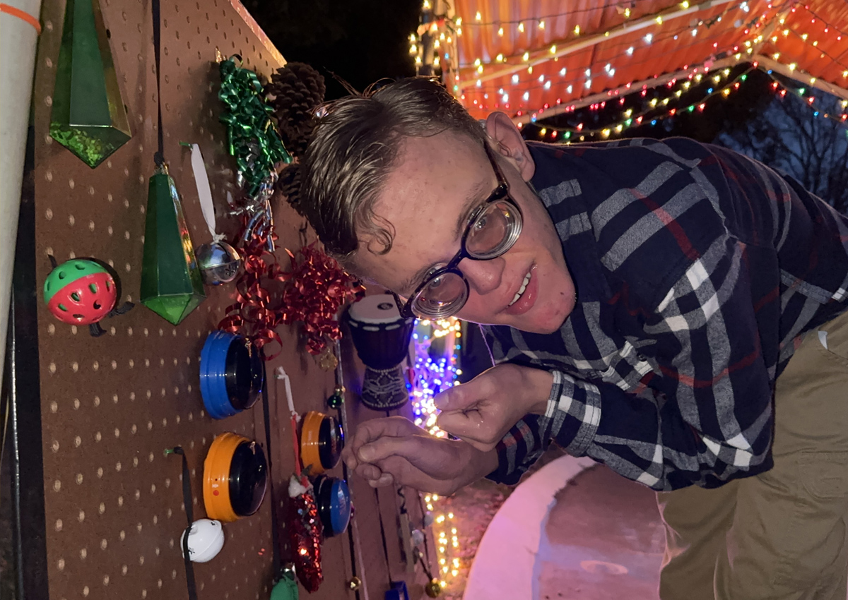 An adolescent crouches next to a peg board with instruments and sound-activated devices attached under a carport covered in Christmas lights in the evening on an outdoor track.