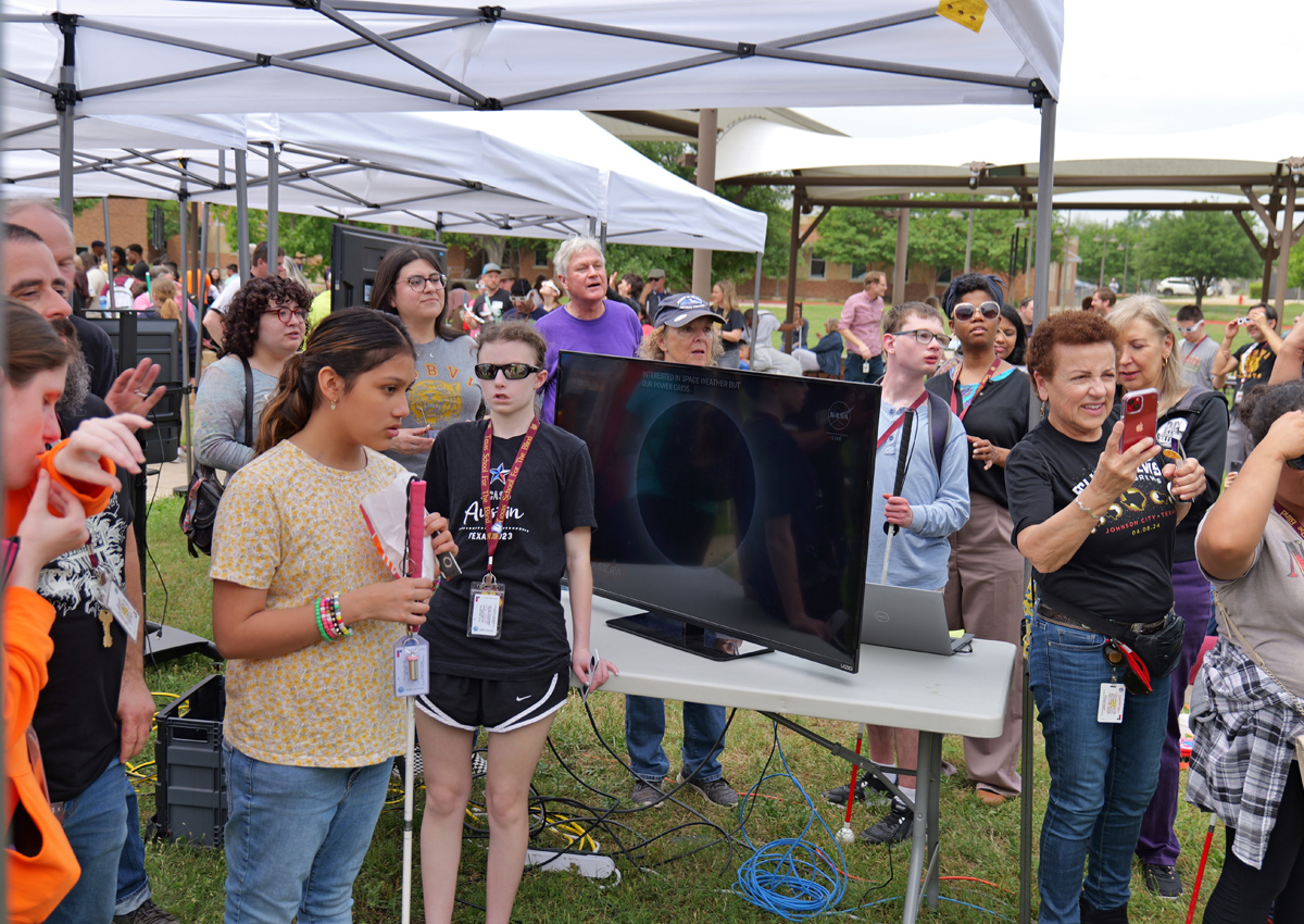 A crowd gathers to experience the eclipse via projected streams on large screens, with LightSound boxes in amplified speakers, and using a telescope with a solar filter and a camera attached to send the images to a computer. From the computer the images were sent to 4 different 46 inch monitors.