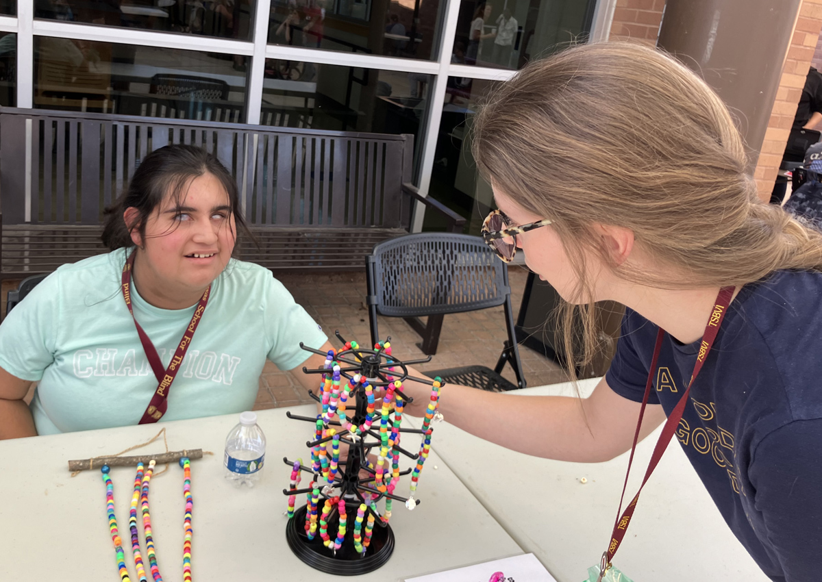 An adolescent sits at a table with beaded bracelets for sale and an adult stands over the rack making a selection.
