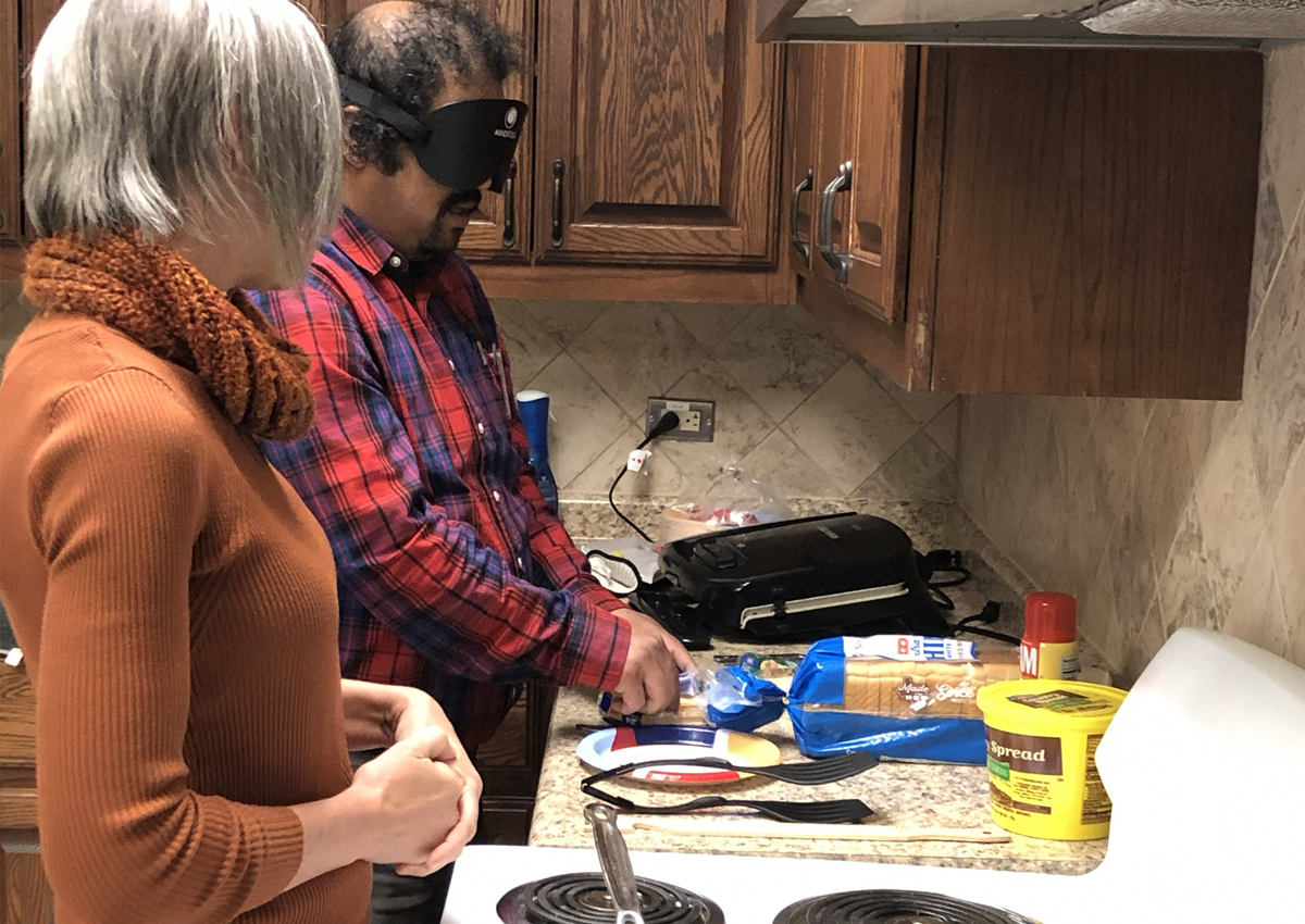 An adult under blindfold prepares a sandwich in a kitchen while an adult watches next to him.