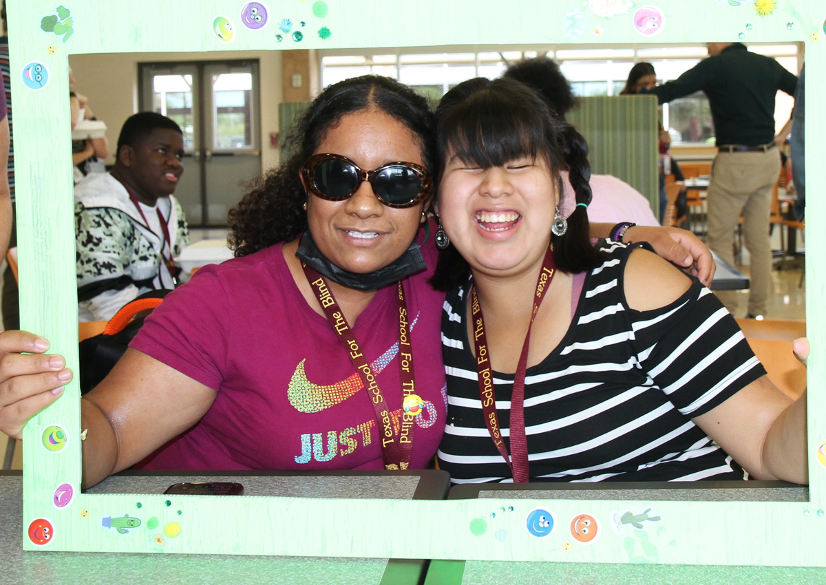 Two adolescents pose close while holding up a cardboard frame seated in a cafeteria.