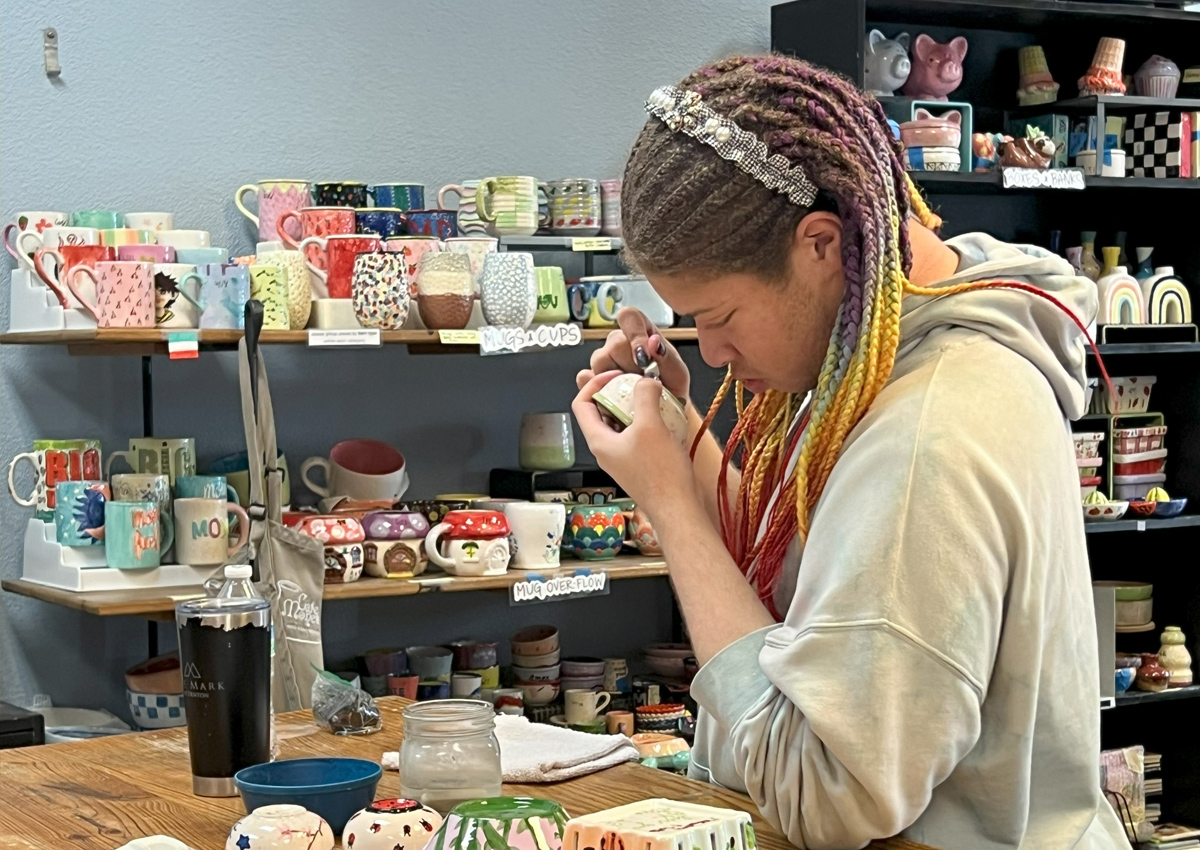 An adolescent uses a tool to mark a slab of clay in a room filled with ceramics.
