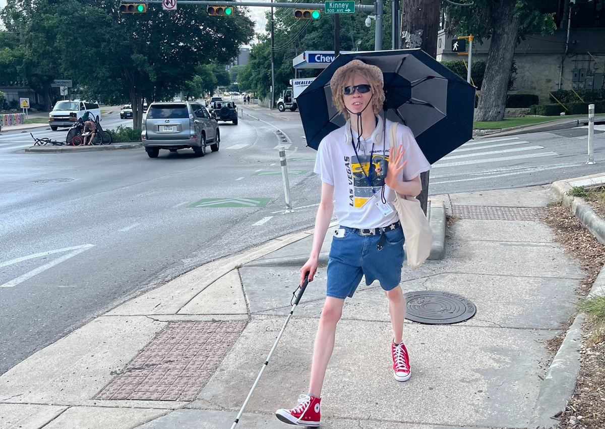 An adolescent uses a straight cane and holds an umbrella as he walks on a city street outdoors.