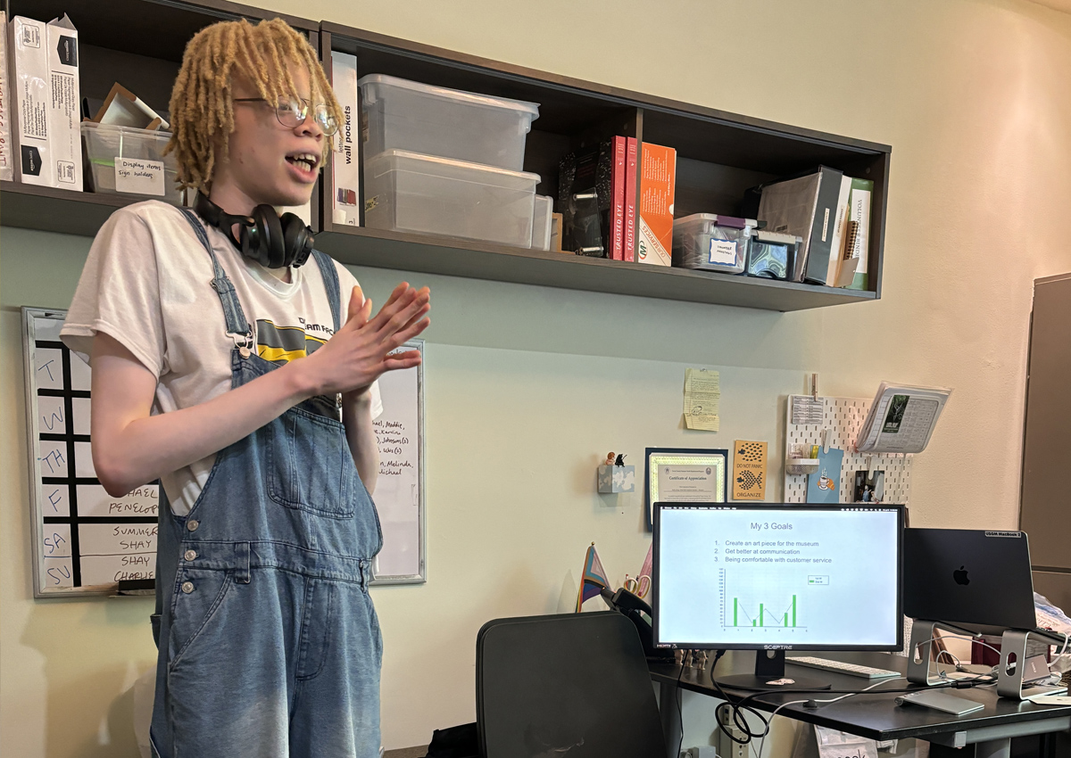 An adolescent stands in an office next to a computer screen with a presentation stating 