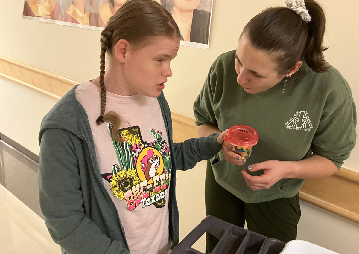 An adolescent pushing a cart in a hallway hands a container of snack mix to an adult.