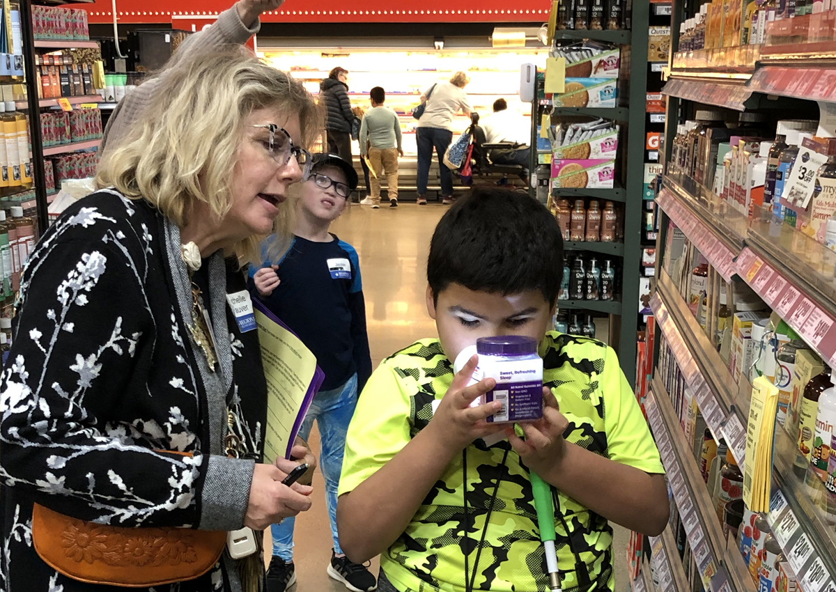 An adolescent holds a magnifier with illumination to a jar of supplements with an adult to his right in the aisle of a grocery store.