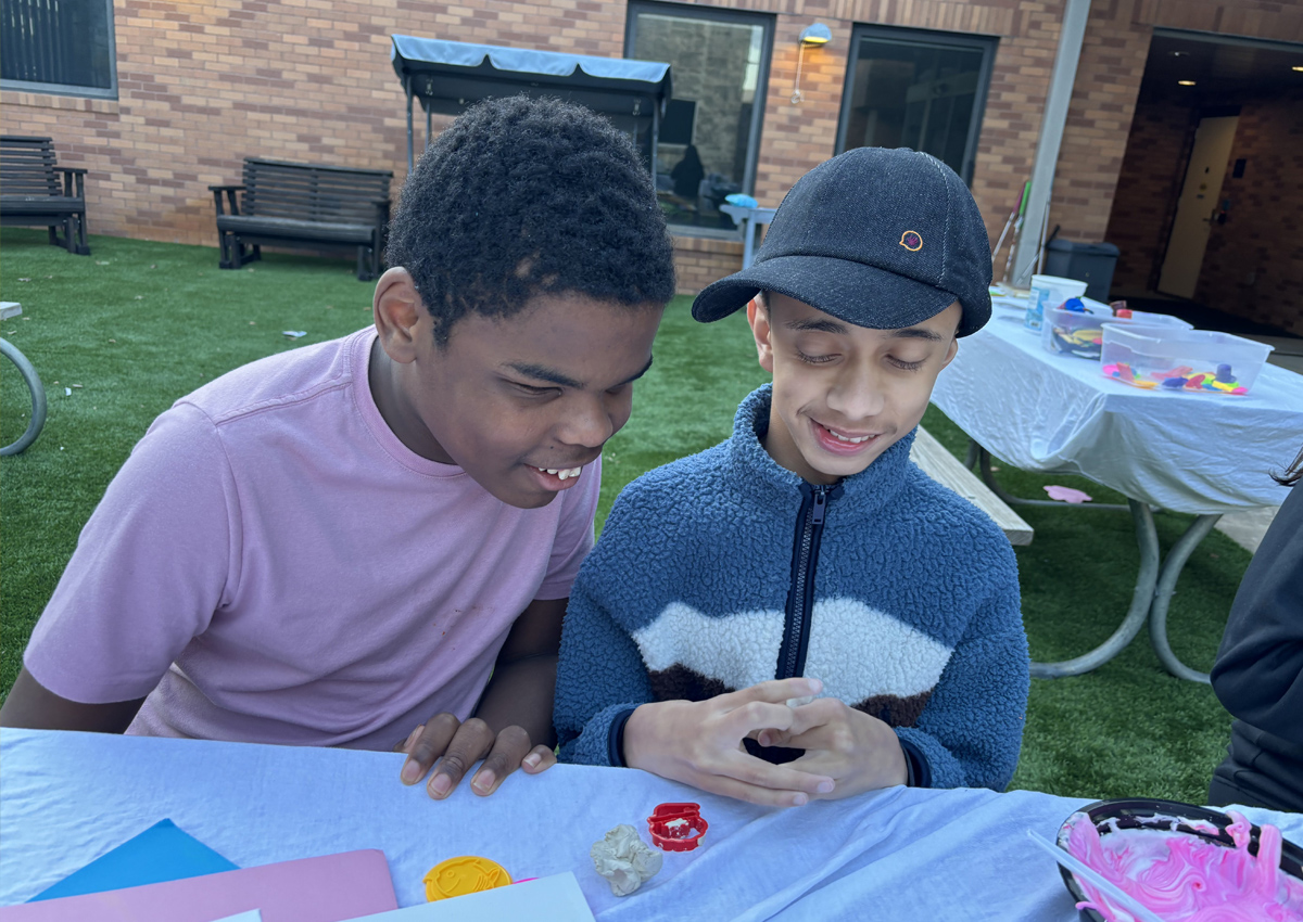 Two adolescents sit outdoors at a picnic table with a bowl of slime on the table.