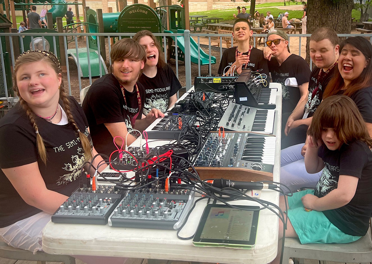 Seven adolescents wearing matching Semi-Modulars shirts sit at a long table with analog synthesizers and digital samplers on an outdoor patio near a playground. An adult crouches next to them.