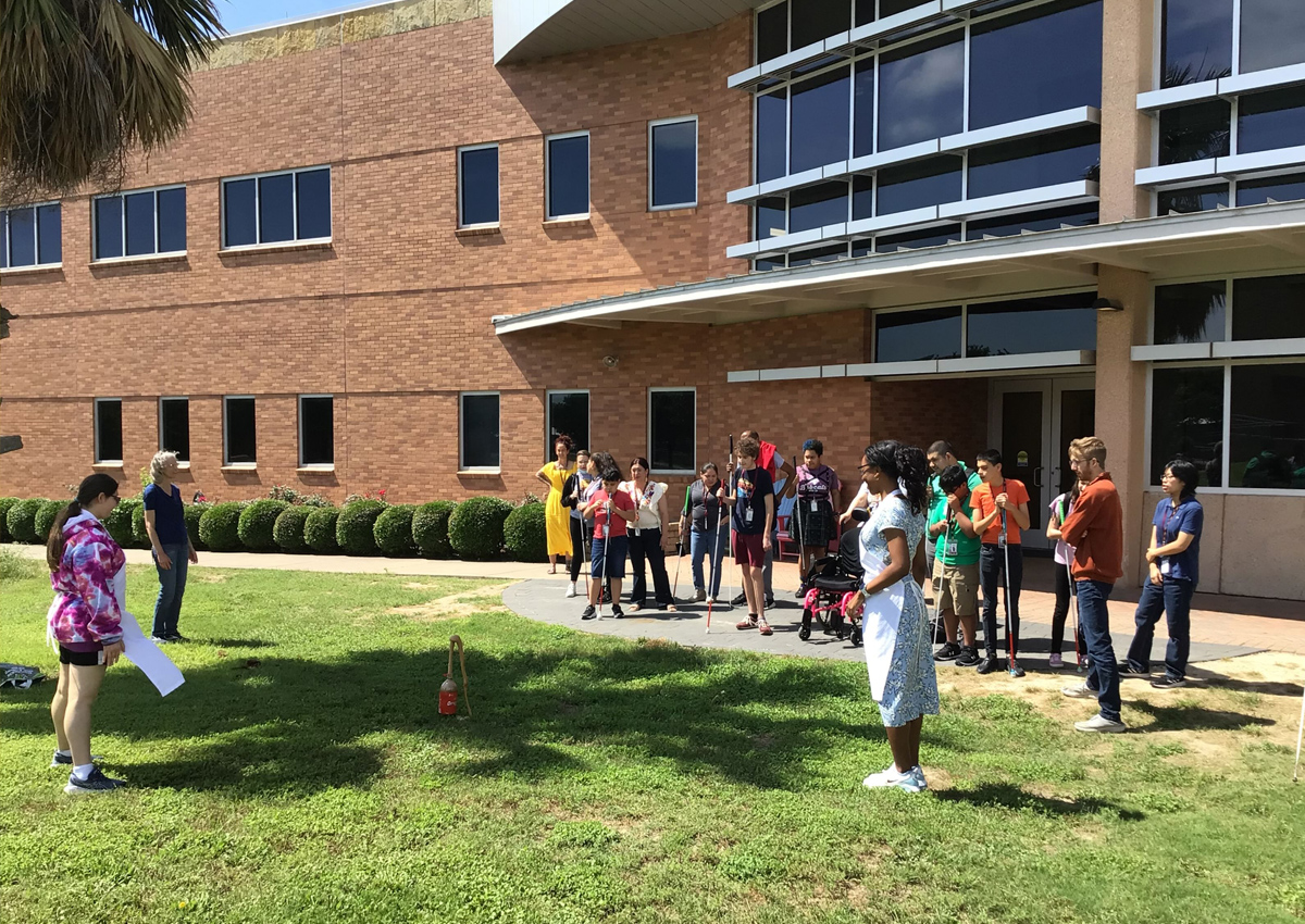 A large group assembles outside a school building for a demonstration in the grass on how to explode coke bottles when Mentos and different ingredients are added.
