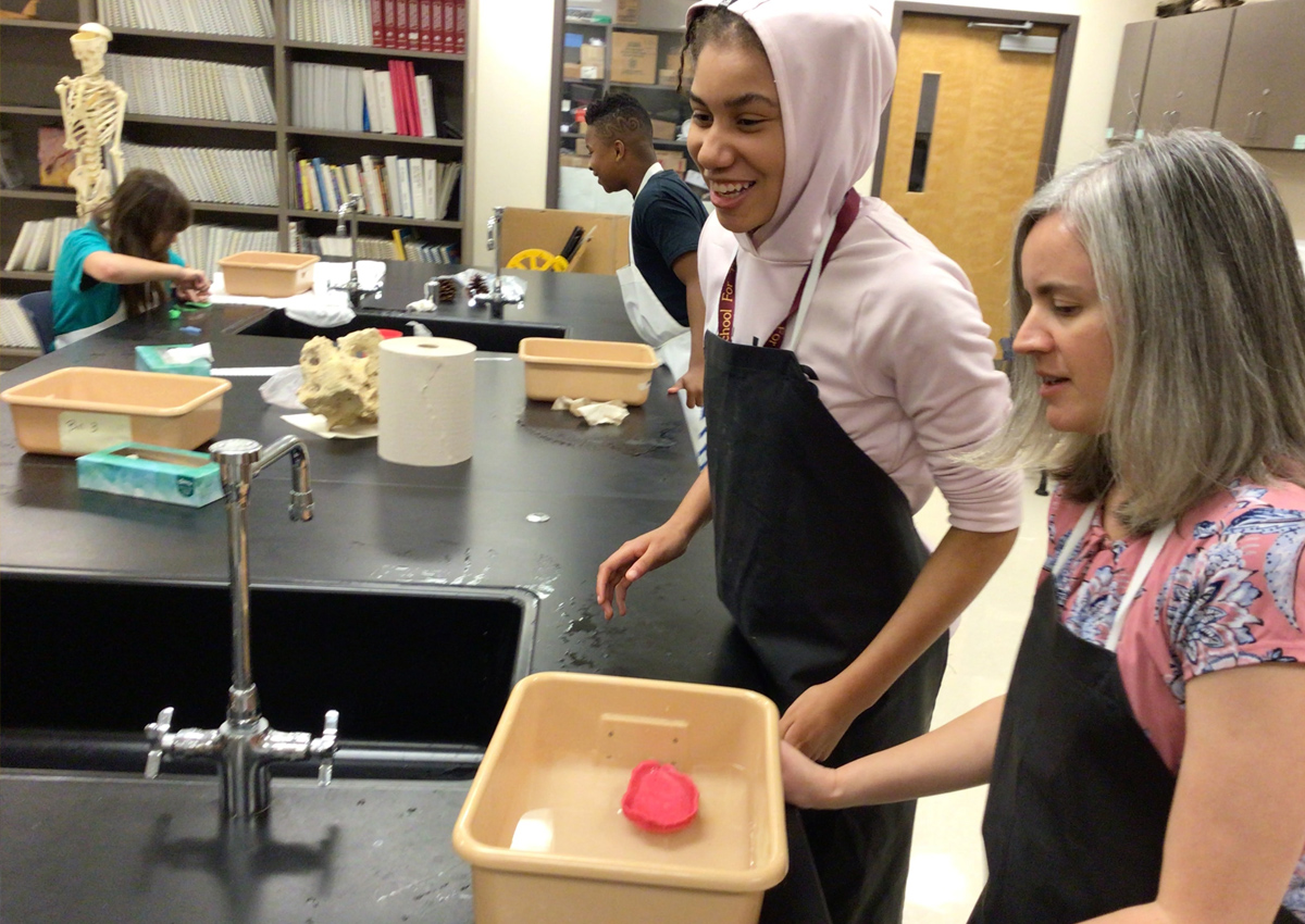 An adult and adolescent stand at a sink in a science lab with a bin of water with objects that have sunk or are floating in the bin.