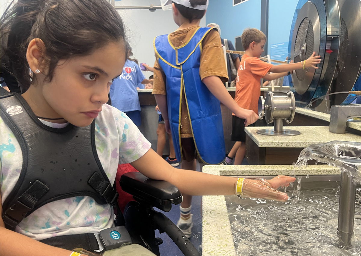 An adolescent in a wheelchair puts her hand in a fountain in an indoor water room at a children's museum.