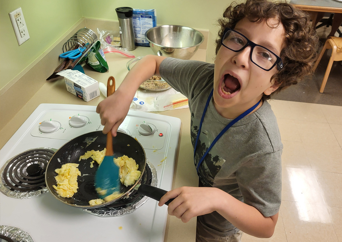 An adolescent smiles while stirring scrambled eggs in a pan on the stove in a kitchen.