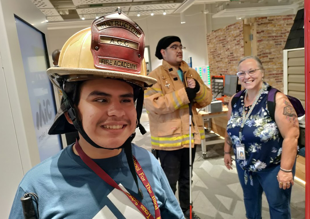 An adolescent wears an official fire academy hat while a peer and adult stand behind him in the distance.