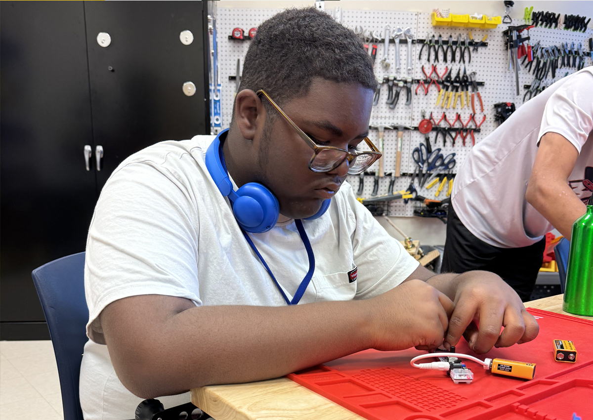 An adolescent works with a small kit of coding wires and batteries on a tray in a makerspace with tools.