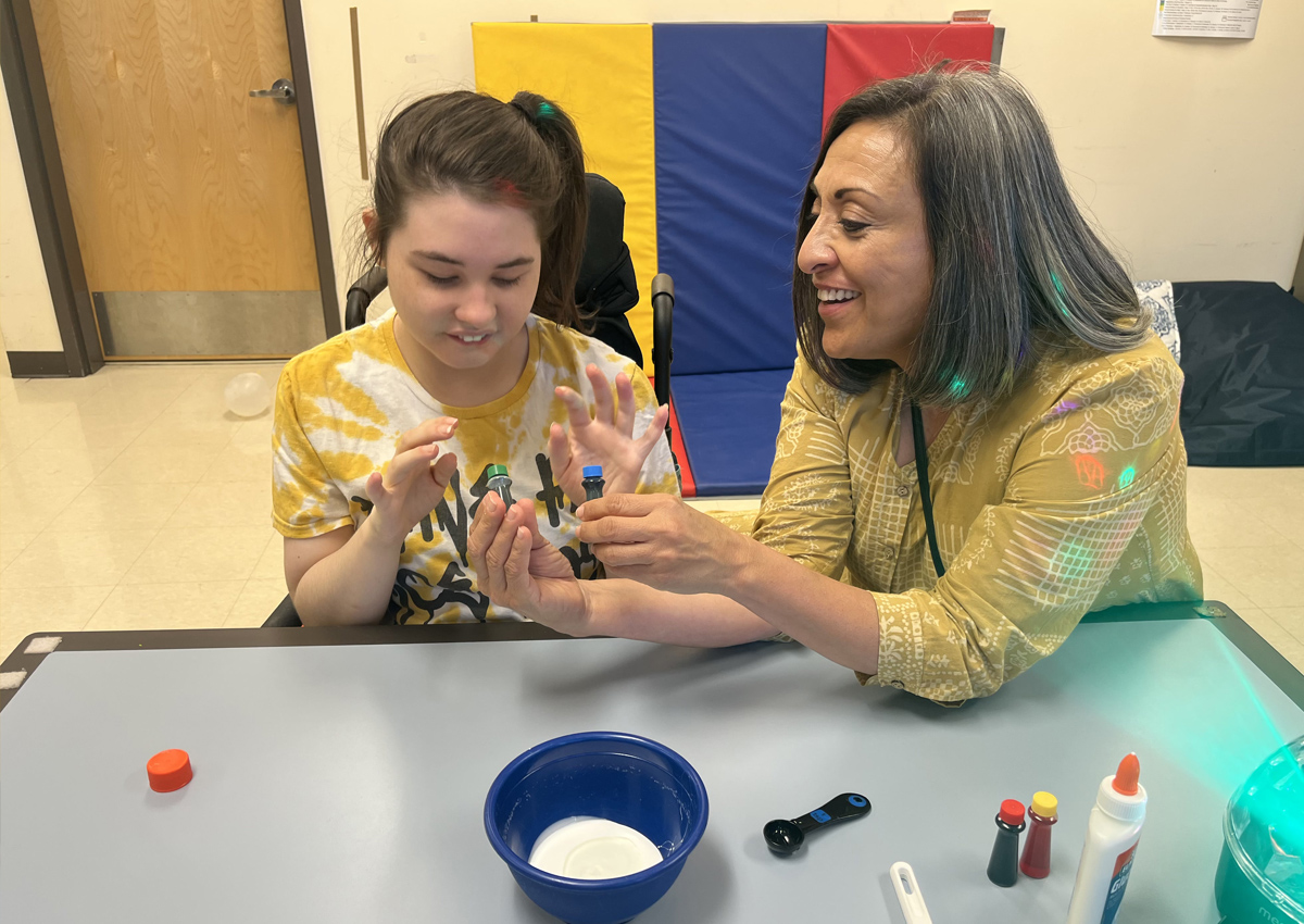An adult presents two choices of food coloring to an adolescent seated at a table. A light projector, bowl of powdery substance, and glue are on the table.