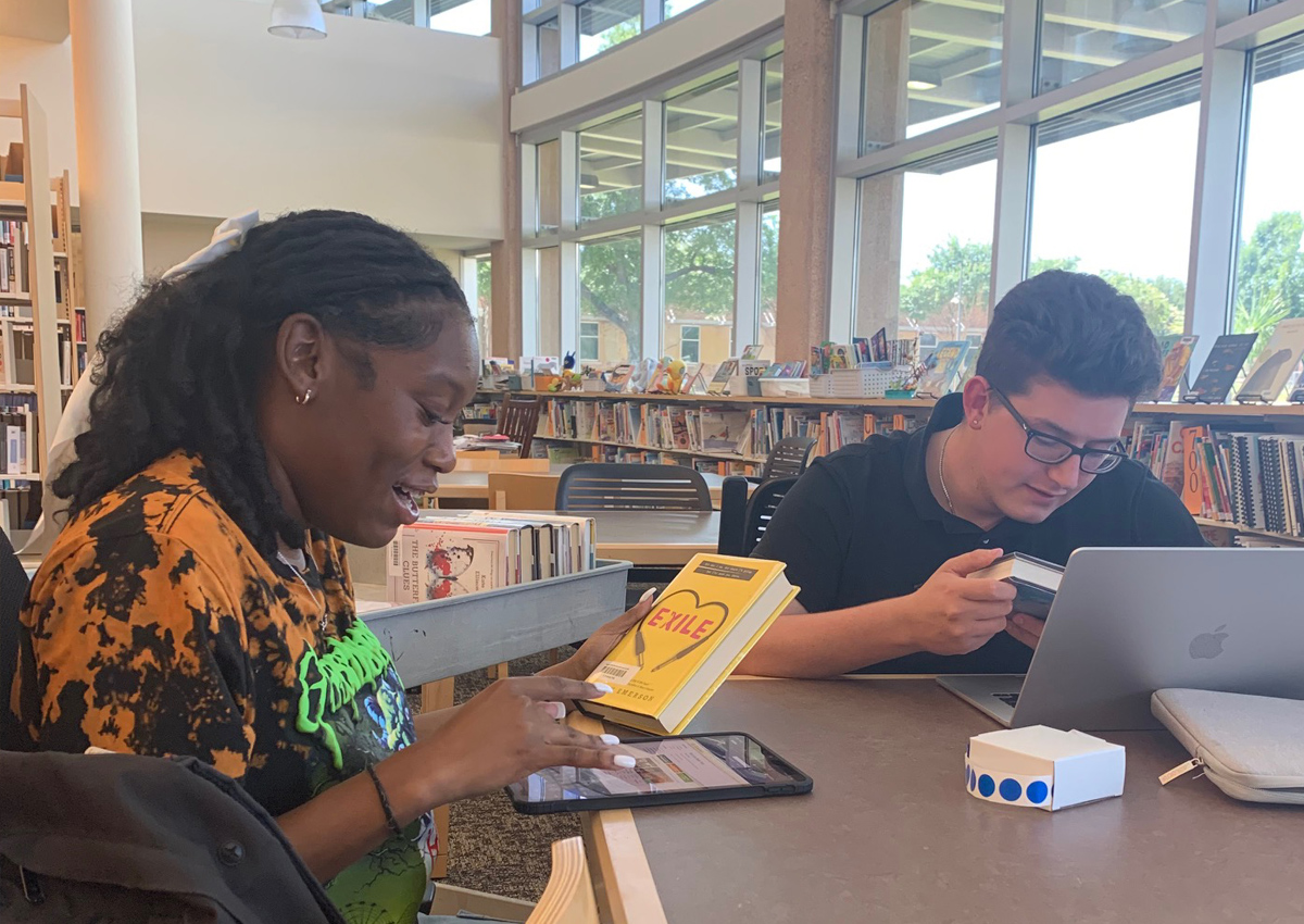 Two adolescents label books in a library using a digital systems on an iPad and laptop.