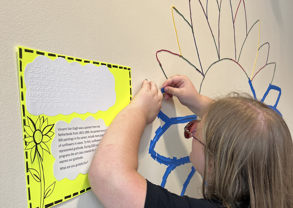 An adolescent puts colorful wiki-sticks on pieces of tape arranged in a large sunflower on a wall. A sign about Vincent Van Gogh in print and braille is on the wall.