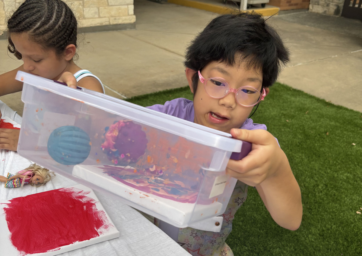 Two adolescents seated at a picnic table outdoors paint by rolling a ball on a canvas in paint in a closed plastic container.
