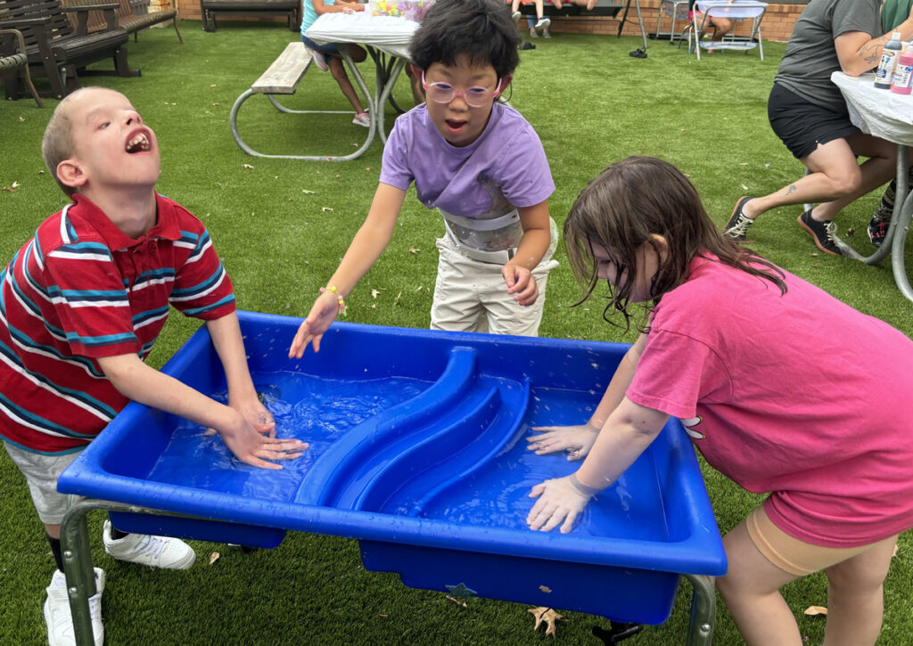 Three adolescents splash in a water table on turf outdoors.