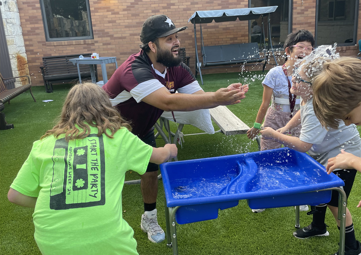 An adult splashes four adolescents from a water table on turf outdoors.
