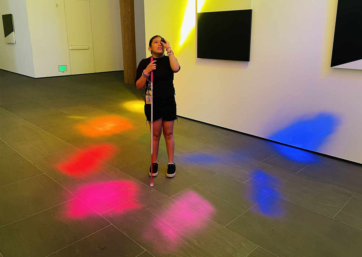 An adolescent with a white cane looking up using a monocular stands in a museum with colored reflections of rainbow light on the floor around her.