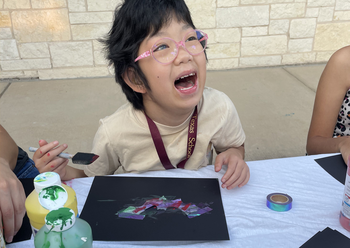 An adolescent seated at an outdoor picnic table smiles as she paints a bat.