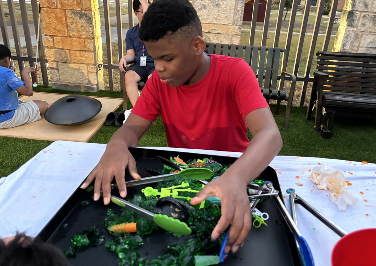 An adolescent seated at a picnic table outdoors searches for Halloween objects in a tray of Jello.