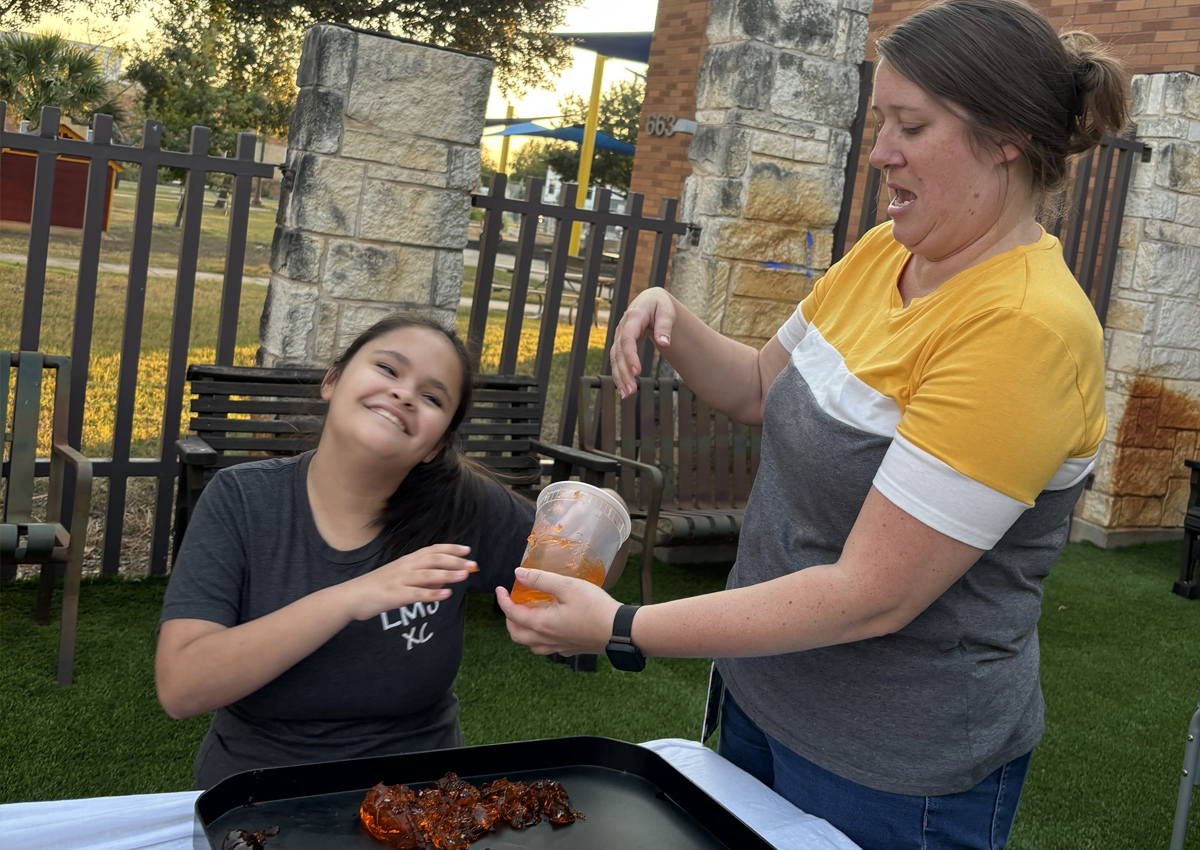 An adolescent seated at a picnic table outdoors rubs her hands in Jello on a tray while an adult standing near holds a container of Jello.