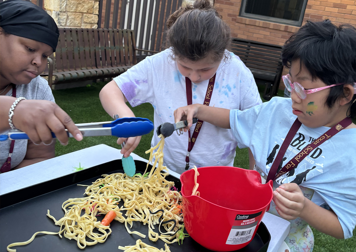 Two adolescents use tongs to find Halloween toys mixed in with long, cooked noodles at a picnic table outdoors.