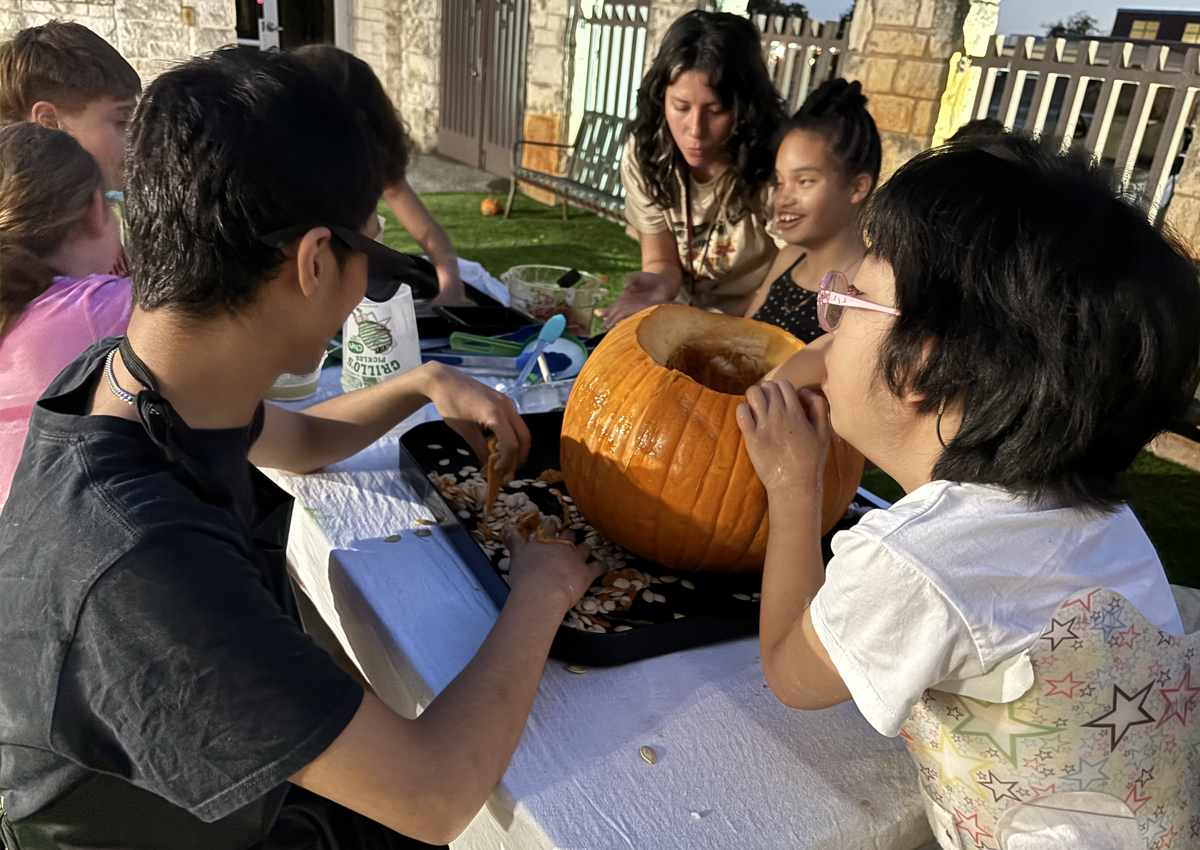 Five adolescents and an adult seated at a picnic table outdoors in the evening carve pumpkins.