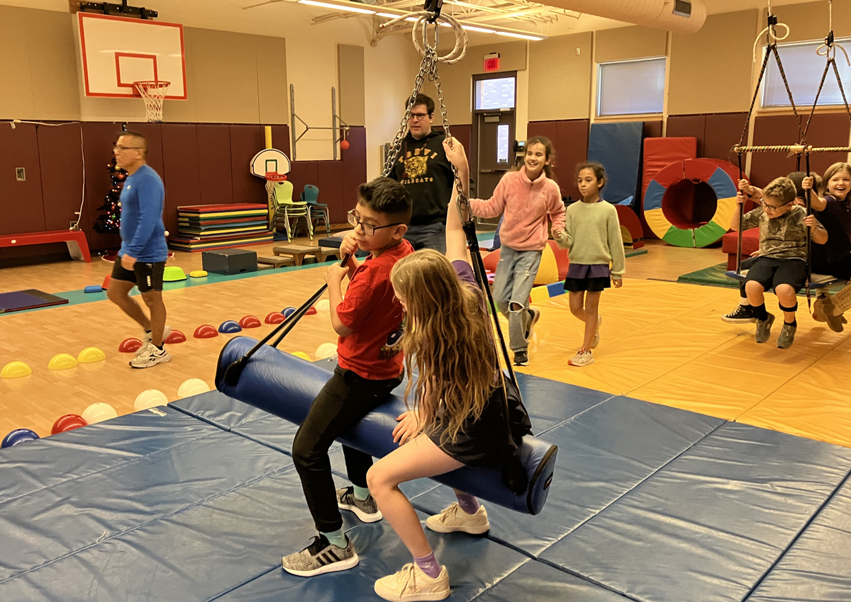 Groups of adolescents engage in PE activities such as swinging on a bolster and flat swing and obstacle course in a gym.