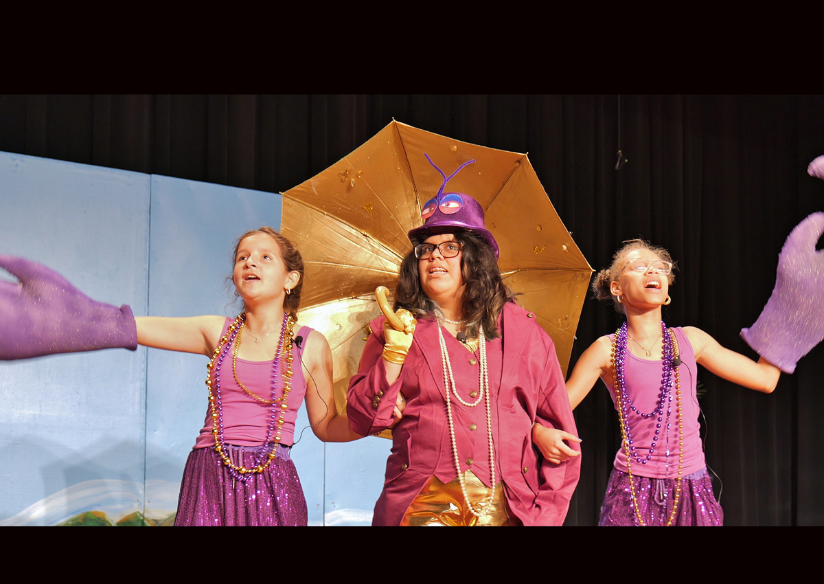 An adolescent holding a gold umbrella with a crab hat and two adolescents each holding a purple crab claw perform 