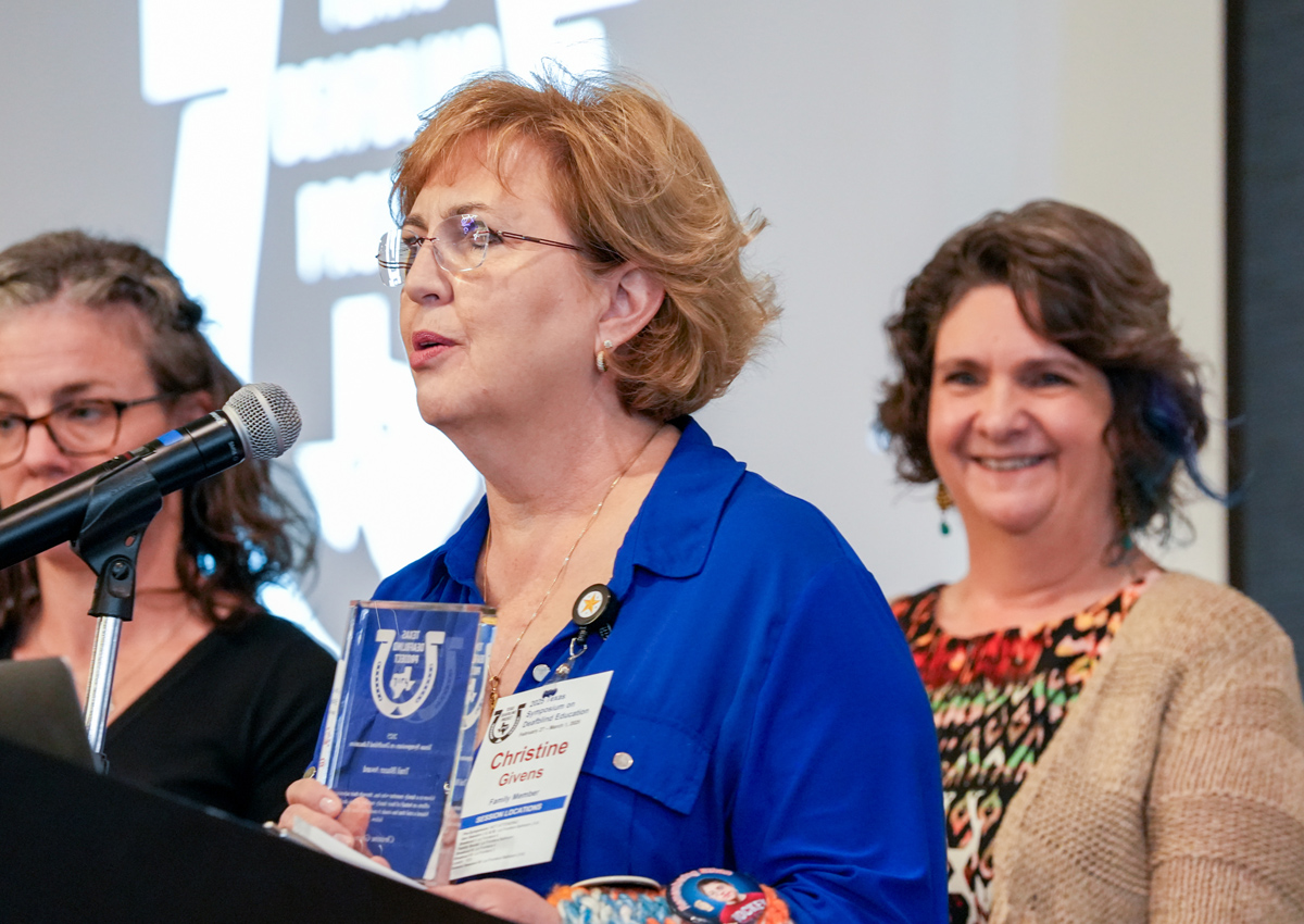 An adult holds a glass plaque and speaks into a microphone at a podium while two adults smile behind her.