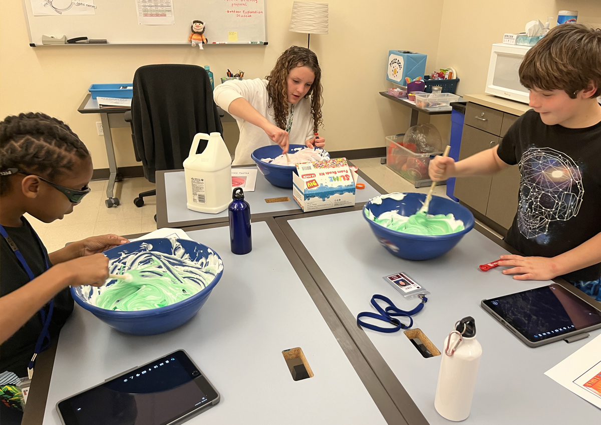 Two elementary students and an adolescent stir slime in large bowls with a large print recipe on the table for each.