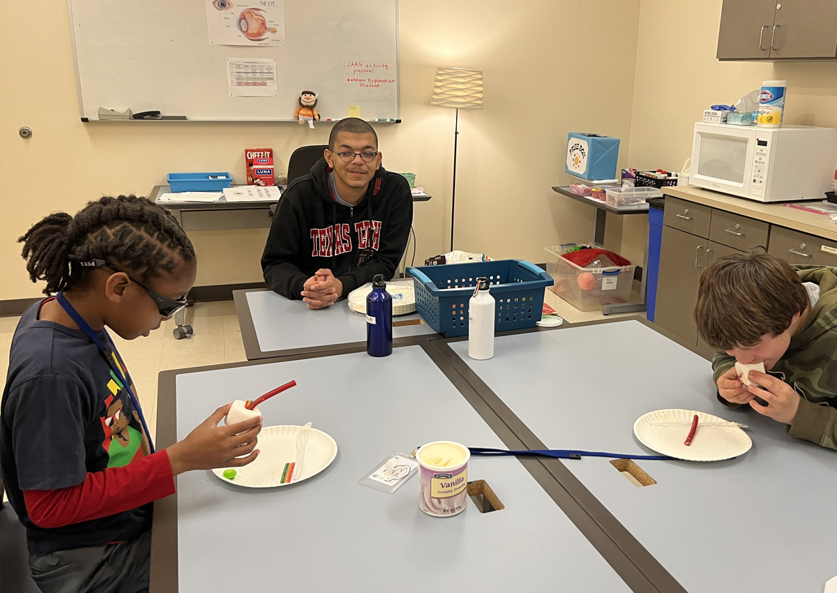 An adolescent and two elementary students at a large table make an edible eye out of a jumbo marshmallow, licorice, frosting, and other ingredients.