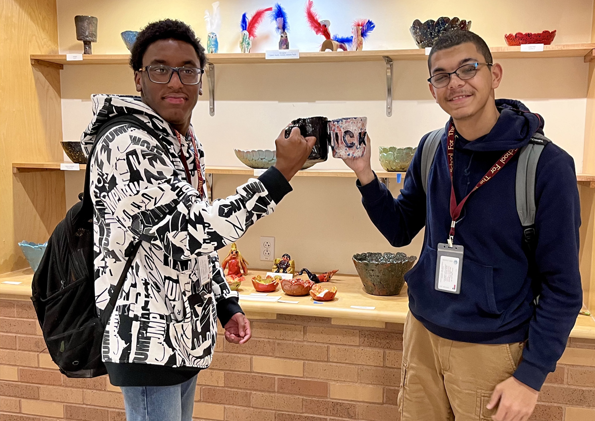 Two adolescents stand in a hallway in front of a display of art and cheers with two homemade ceramic mugs.