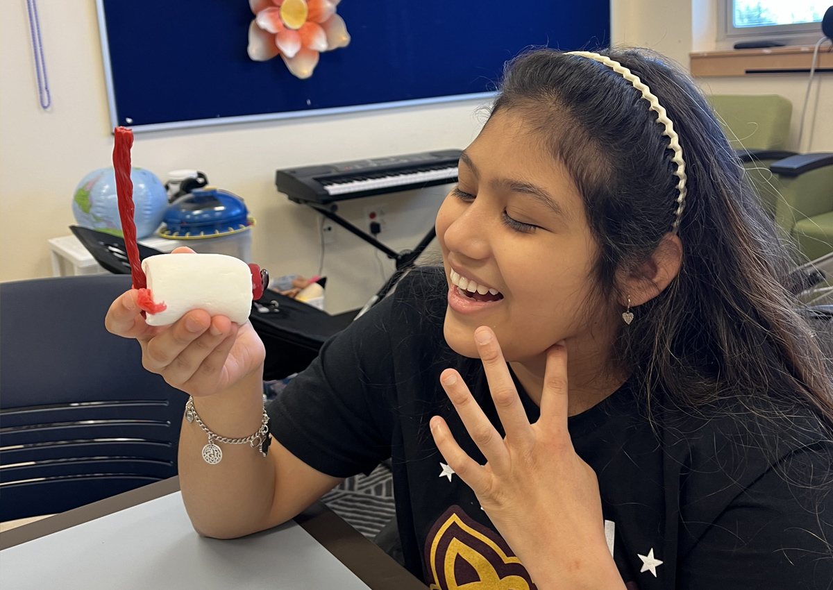 An adolescent smiles as she holds up the edible eyeball she made out of candy (licorice, jumbo marshmallow, lifesaver, gum drop).