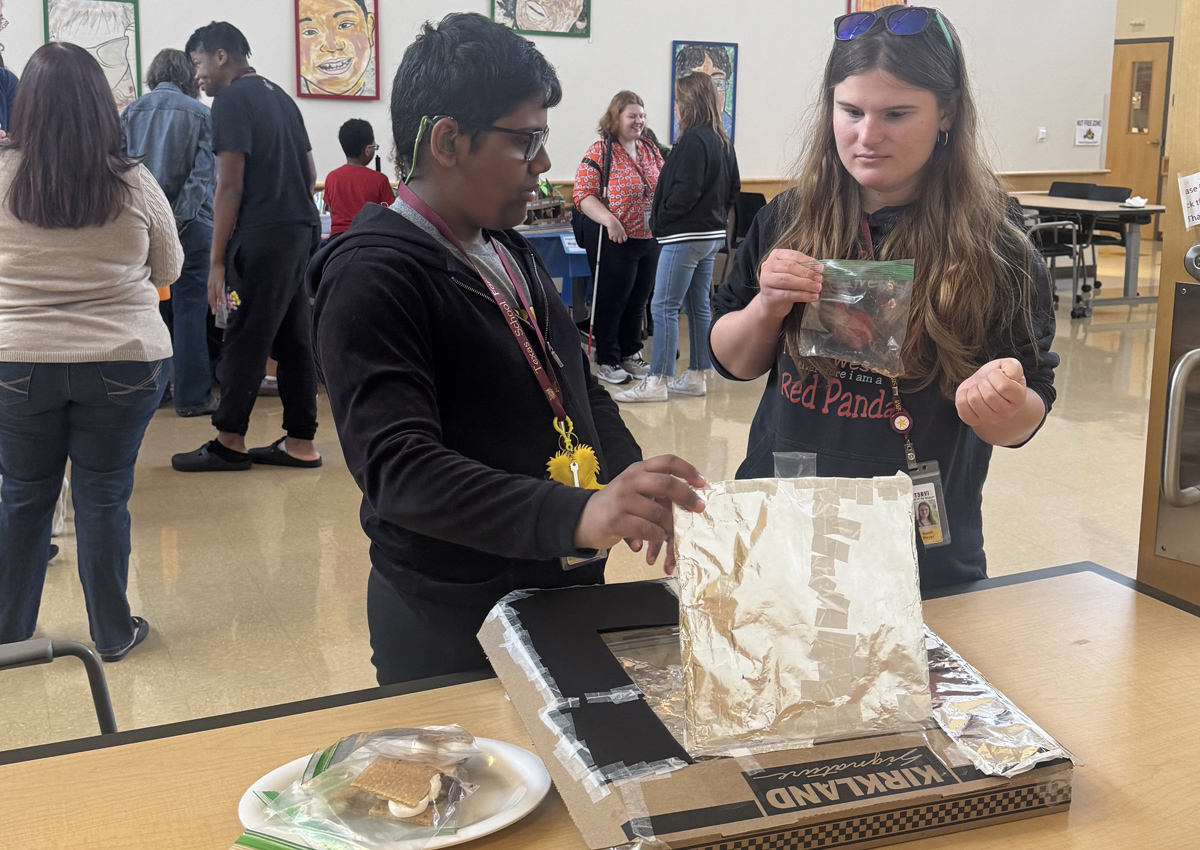 An adolescent demonstrates a solar oven made from a large flat pizza box, aluminum foil, and packing peanuts with bags of S'mores next to the oven and given to a peer.