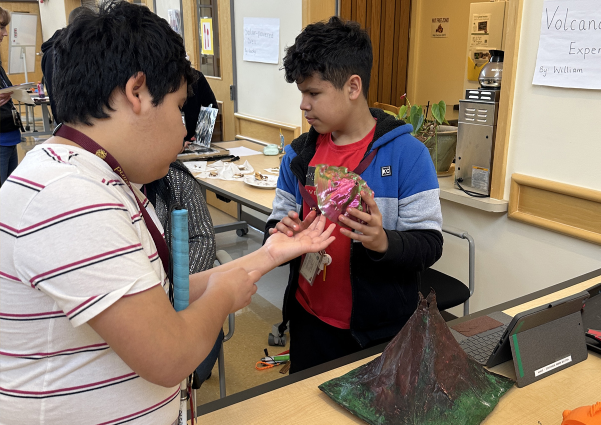 An adolescent shows a 3D printed volcano to a peer while a larger model of a volcano and an iPad rest on the table nearby. A sign reads 
