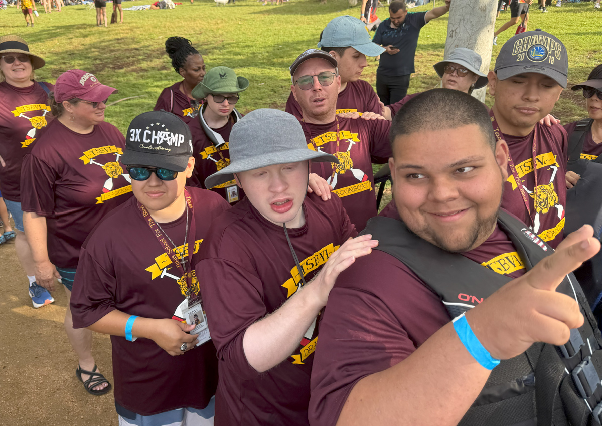 A group of adolescents wearing matching TSBVI Dragonboat shirts line up behind each other at a park.
