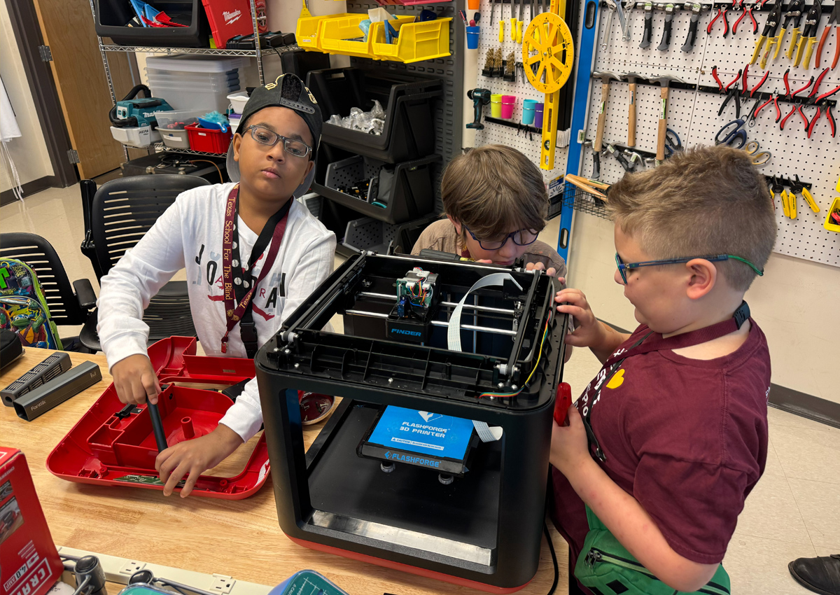 Three adolescents take apart a large 3D printer using tools. Tools hang on peg boards. on the wall