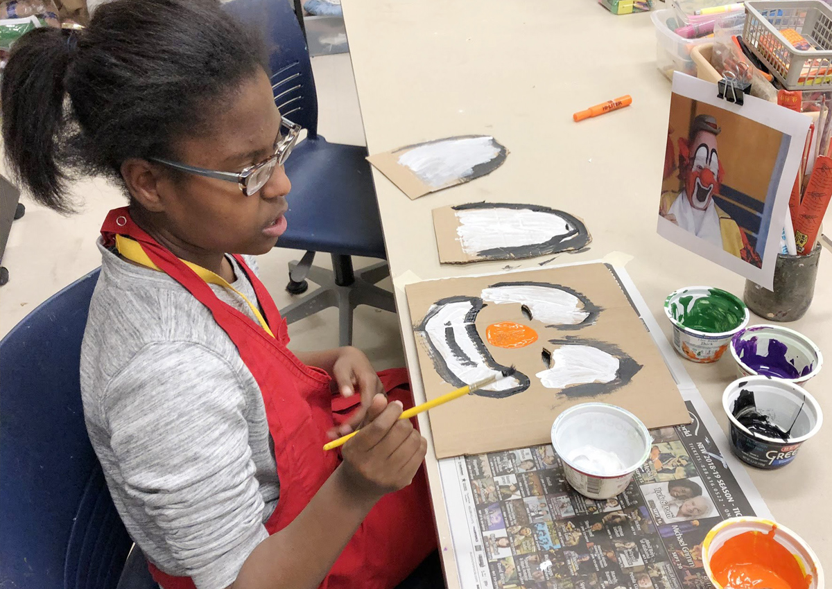 An adolescent in an apron sits at an art table painting on a clown face. A book holding a picture of a clown stands upright as a model. Five containers filled with paint are on the table.