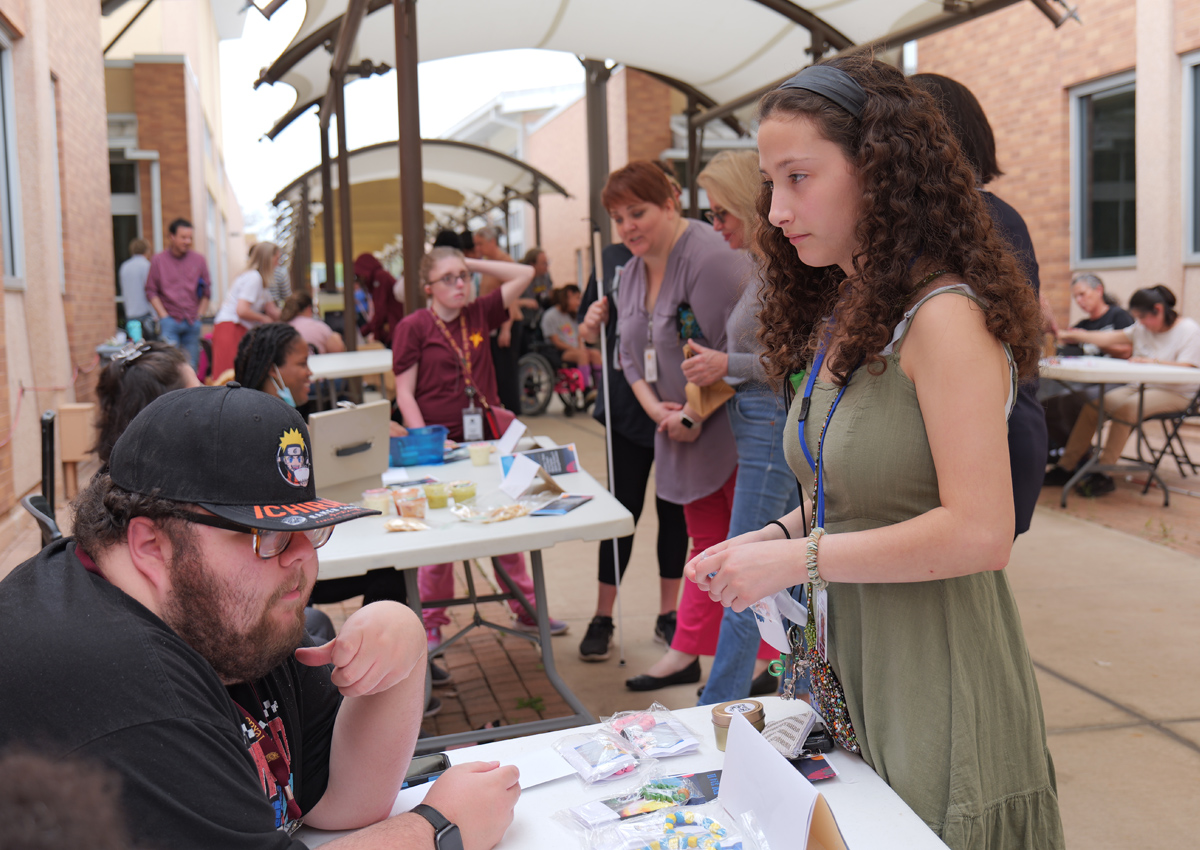 An adolescent buys a product from another adolescent seated at a table in an open market.